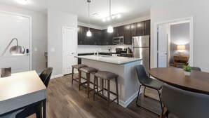 A modern kitchen with dark cabinets, stainless steel appliances, a central island with stools, and a dining area with a table and chairs. - Carolina Forest Furnished Apartments by Landing  (Myrtle Beach)