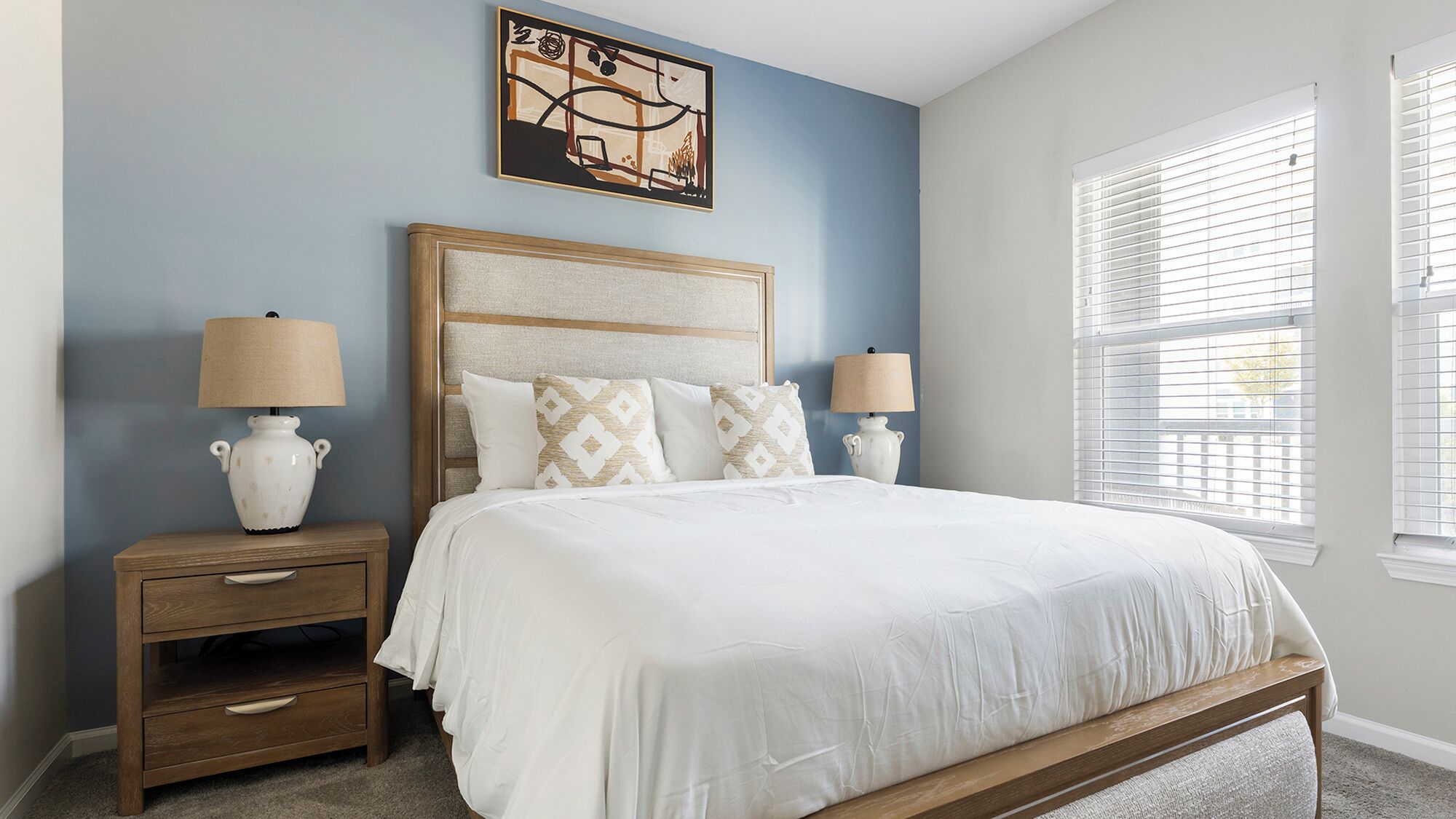 A bedroom with a large bed, two bedside tables with lamps, a decorative wall piece, and a window with blinds.