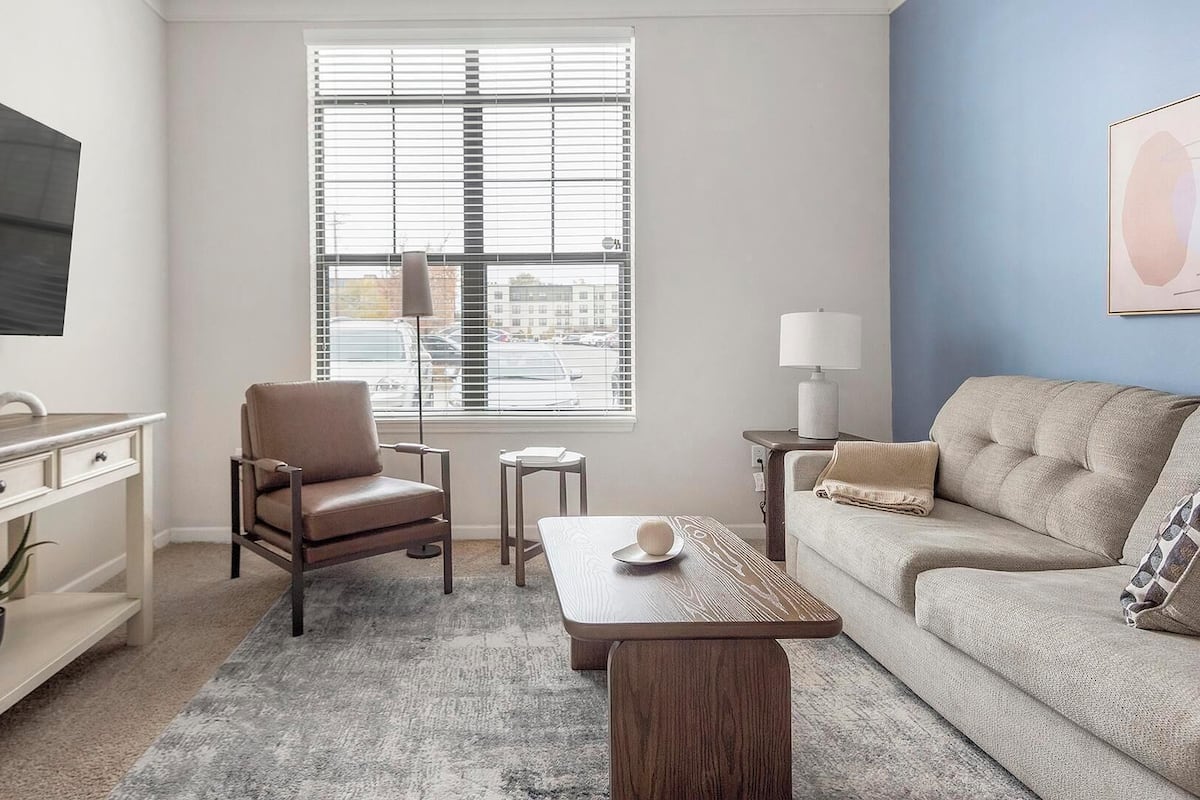 Un salon moderne avec un téléviseur à écran plat, un fauteuil en cuir, une table basse en bois, un canapé beige et une fenêtre avec des stores.