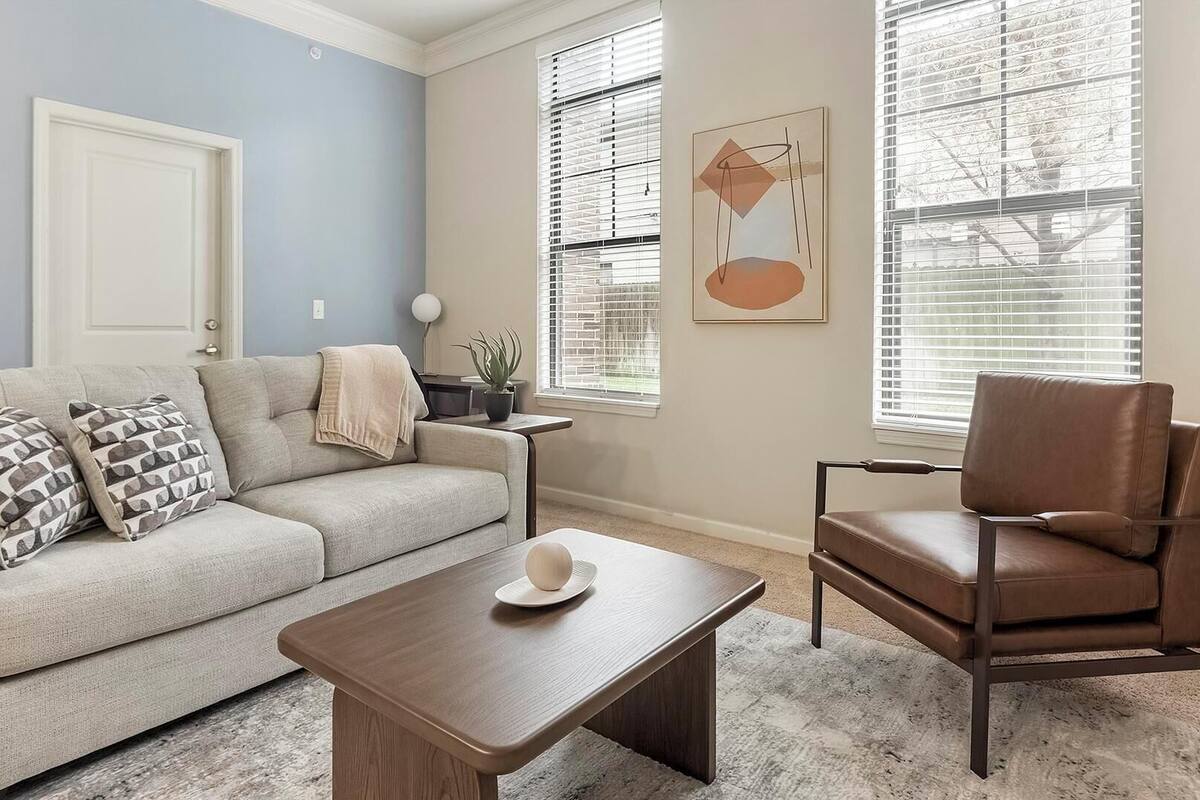 Un salon comprenant un canapé, une table basse et un fauteuil. On y trouve une porte, une fenêtre avec des stores et une œuvre d’art accrochée au mur.