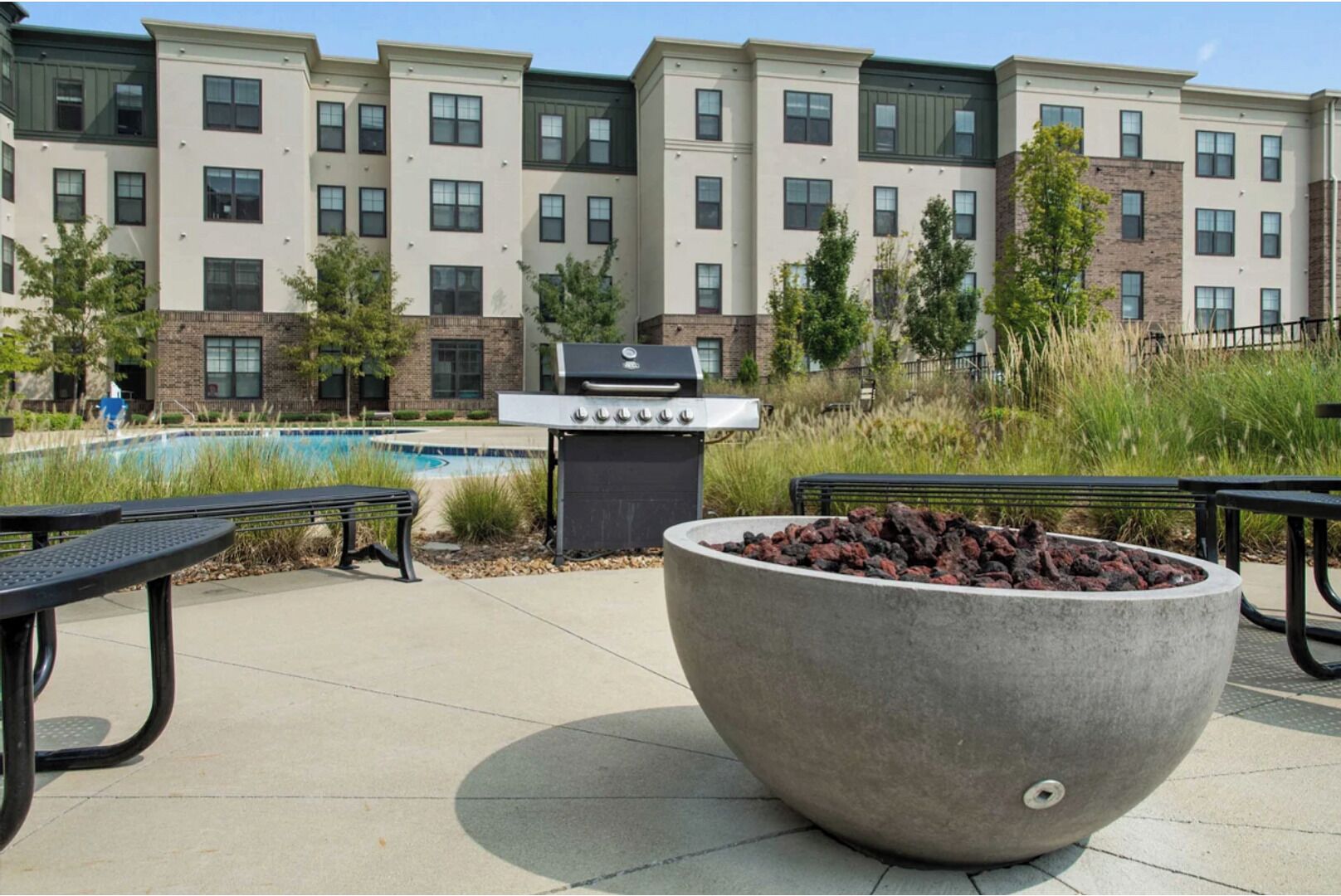 A patio area with a barbecue grill, a large fire pit, and a pool in the background.