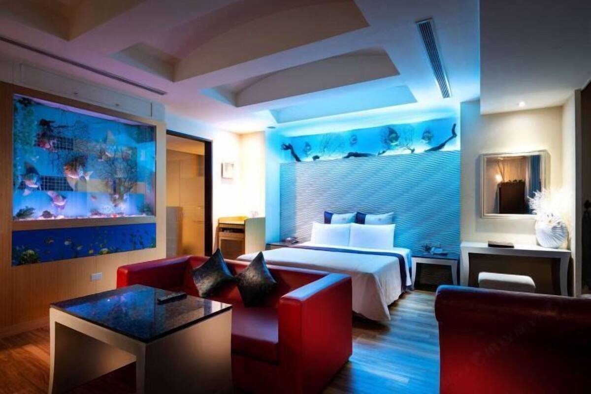 Une chambre d’hôtel avec un lit, un bureau, un canapé rouge et un grand aquarium.