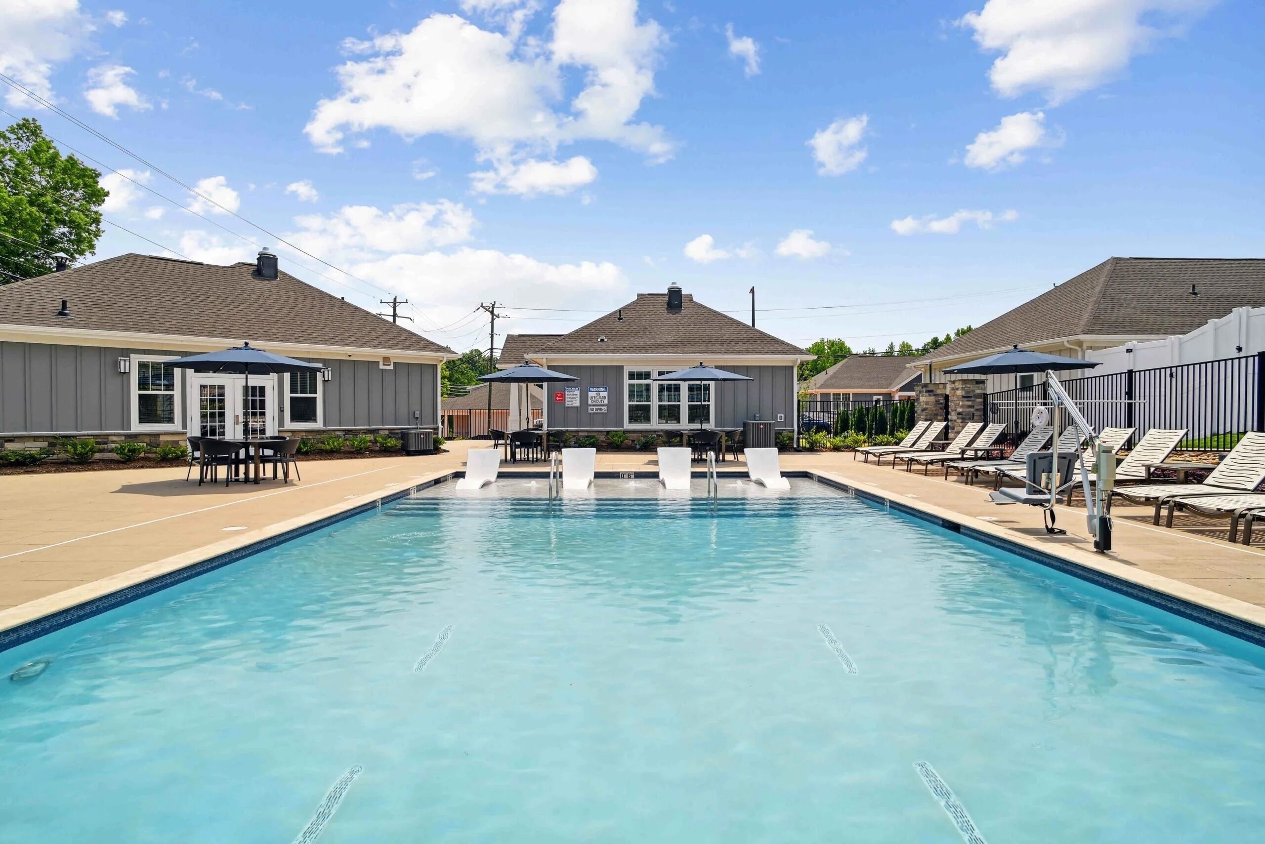 Une piscine entourée de chaises longues et de parasols, elle-même bordée de bâtiments aux façades grises et aux toits en pente.