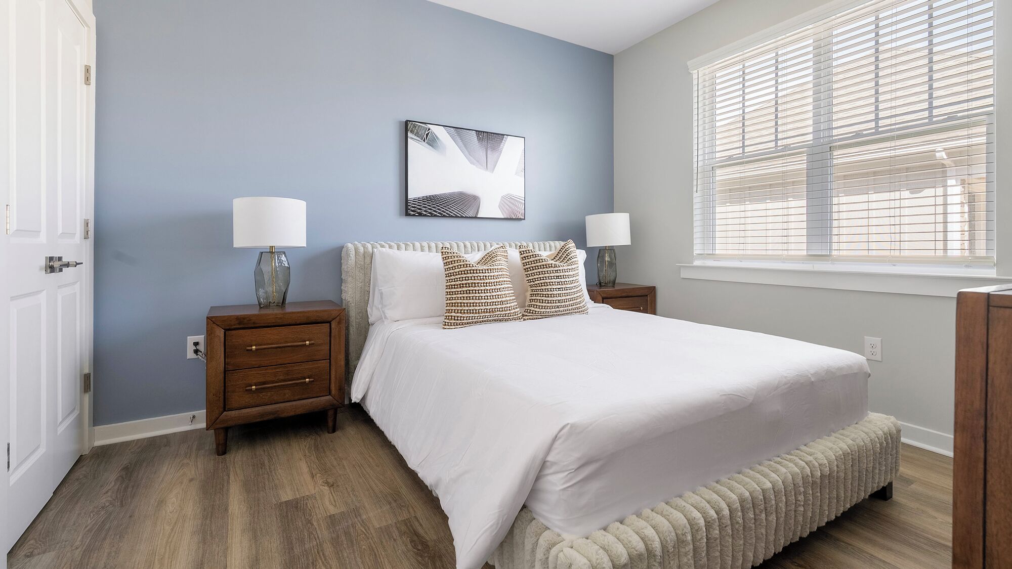 A bedroom with a bed, two nightstands, a wall-mounted lamp, a window with blinds, and a radiator.