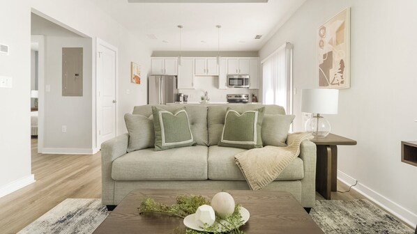 Ein Wohnzimmer mit einer Couch, einem Couchtisch mit Dekoration, einer Küche im Hintergrund und einem Bild an der Wand.