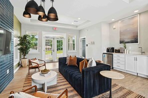 A modern lobby with a dark blue sofa, a round coffee table, and a small round table. There is a potted plant, a large window, and a kitchen area with white cabinets and a sink. - Newell Furnished Apartments by Landing (Charlotte)