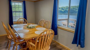 A dining area with a wooden table and chairs, a window with a view of a dock and water, and blue curtains. - Lookout Lodge | Waterfront | Private Dock (Hammond)