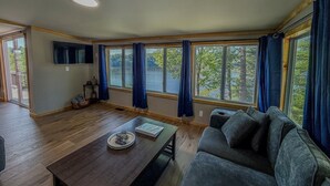 A spacious living room with a large window, a flat-screen TV, a wooden coffee table, and a gray sofa. - Lookout Lodge | Waterfront | Private Dock (Hammond)