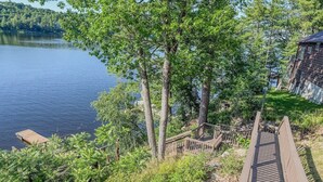 A wooden deck leading to a dock over a lake, surrounded by trees and a small building. - Lookout Lodge | Waterfront | Private Dock (Hammond)