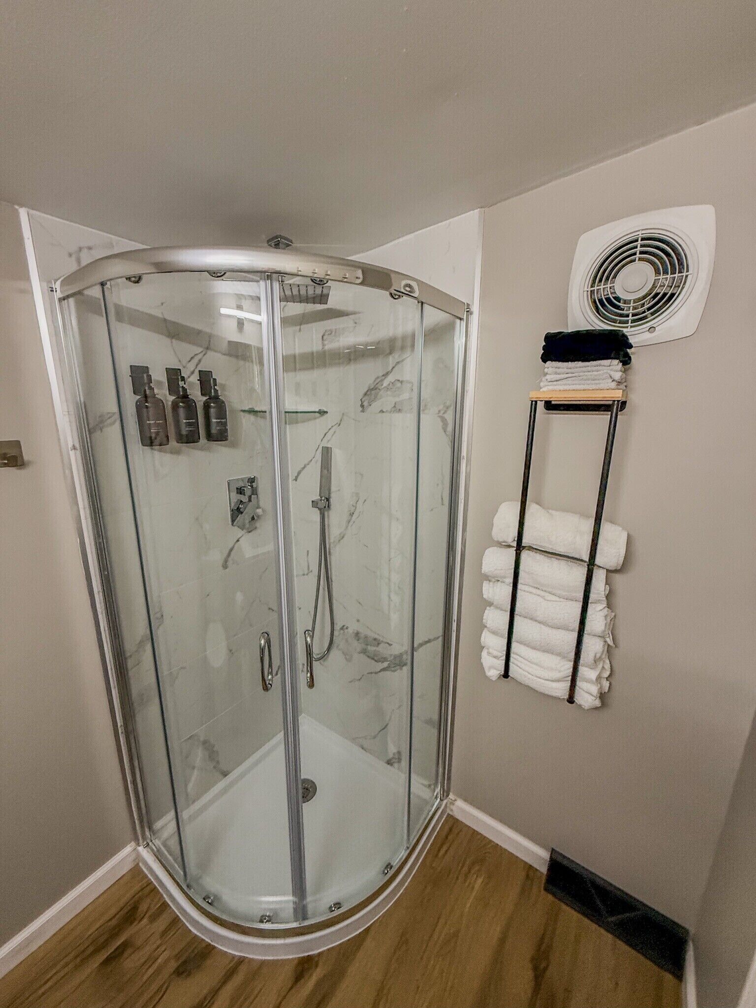 A corner shower with glass doors, a towel rack with folded towels, and a wall-mounted fan.