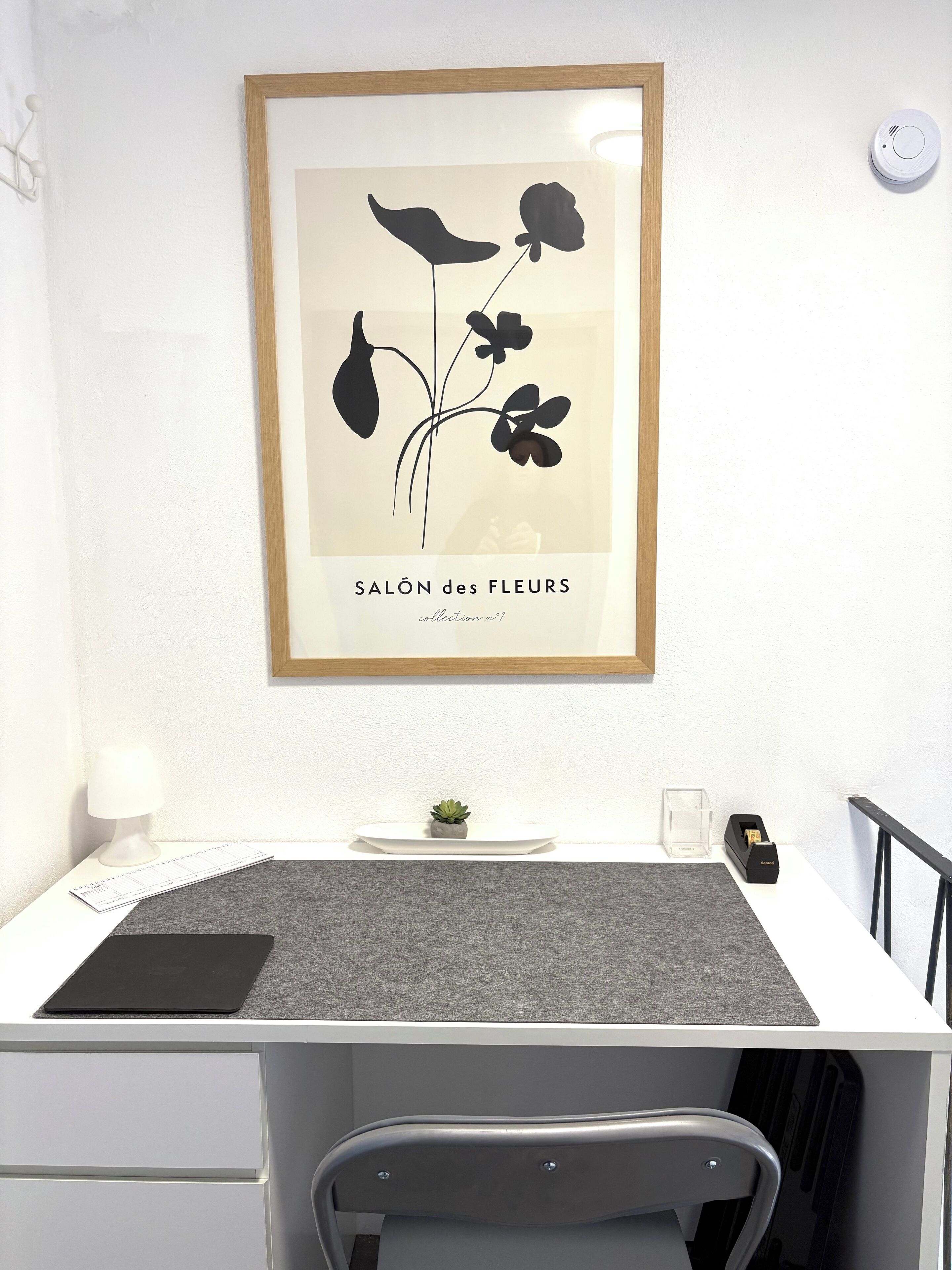 A desk with a framed botanical print, a small potted plant, a desk lamp, and a calendar.