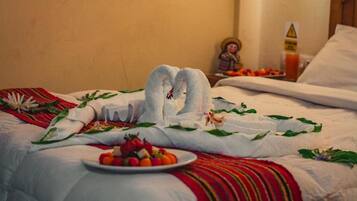 A neatly made bed with a heart-shaped towel arrangement, a plate of fruit, and a small figurine.