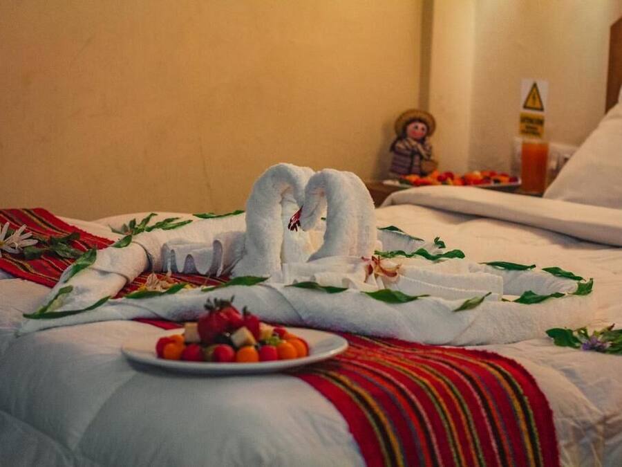 Una cama bien hecha con una toalla dispuesta en forma de corazón, un plato de fruta y una pequeña figurita.