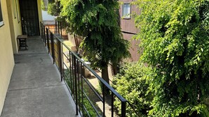 Un balcon doté d’un sol en béton et d’une rambarde en métal, donnant sur un jardin avec des arbres et un bâtiment avec des fenêtres.