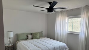 Une chambre à coucher avec un lit, un ventilateur de plafond, une fenêtre avec des rideaux et une lampe de chevet.