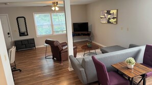 Un salon moderne avec un canapé gris, un fauteuil en cuir marron, une table basse en bois et un téléviseur posé sur un meuble.
