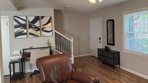 Un salon spacieux doté d’un ventilateur de plafond, d’un fauteuil en cuir marron, d’un bureau avec une imprimante, d’œuvres d’art abstraites accrochées au mur, d’un escalier et d’une fenêtre avec des stores.