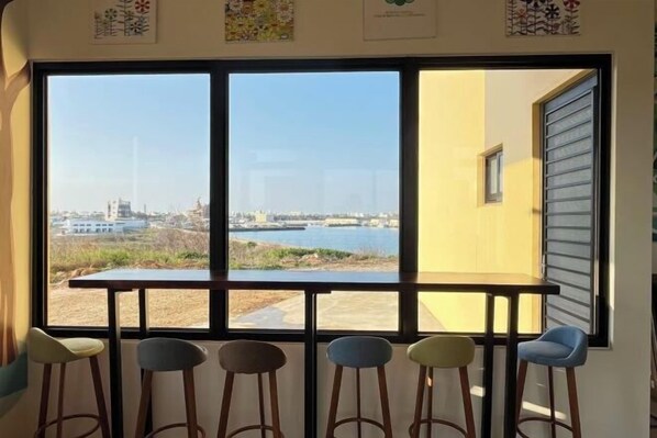 A room with a large window offering a view of a body of water and distant buildings. There are five stools arranged in front of the window. - Su Ye Mu Mu (Penghu)
