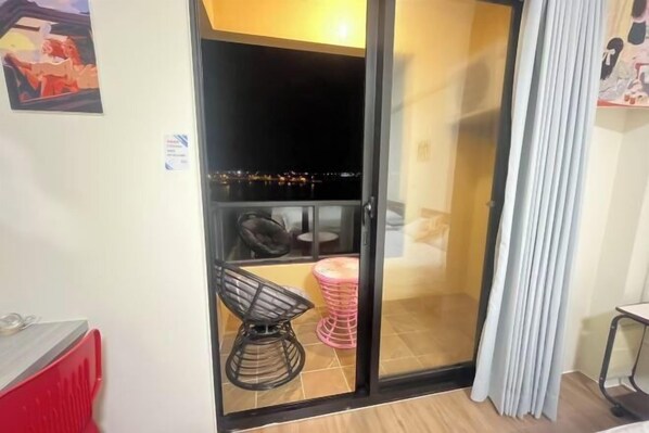 Une pièce dotée d’une porte-fenêtre coulissante donnant sur un balcon, d’un radiateur rouge, d’une chaise noire, d’un tabouret rose et d’un rideau blanc.