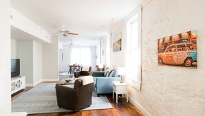 Ein geräumiger Wohnbereich mit einem blauen Sofa, einem Couchtisch und einem Fernseher auf einer Kommode. An der Wand hängt ein Gemälde, und eine Deckenlampe sorgt für Luftzirkulation.