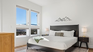 Une chambre avec un grand lit, deux tables de chevet, une œuvre d’art accrochée au mur et une vue sur la ville par les fenêtres.