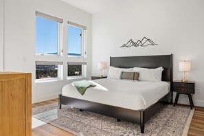 A bedroom with a large bed, two bedside tables, a wall-mounted artwork, and a view of the city through the windows. - Downtown WP Chalet 50 I Hot Tub I Great Views I Car Charger I Bunks I Discounted Attractions (Winter Park)