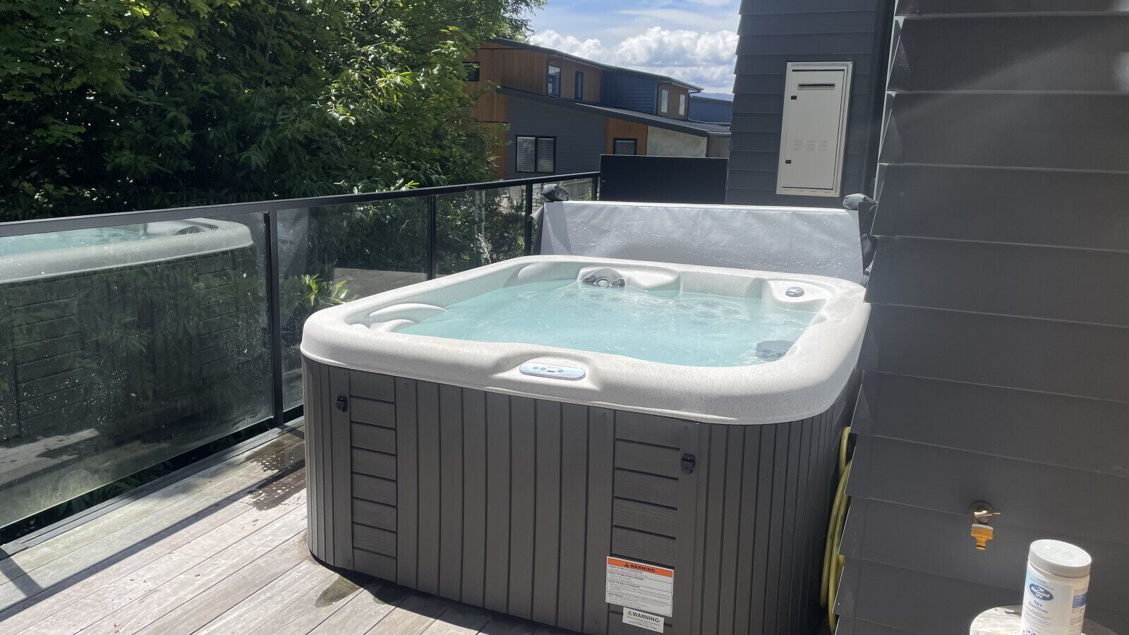A hot tub on a wooden deck with a glass barrier, a metal railing, and a building in the background.