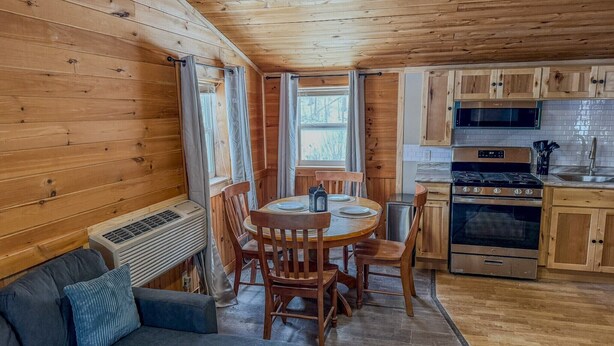 A cozy cabin interior with a dining area, wooden walls, a kitchenette, and a sofa. - Pine & Paddle | Family Cabin | Sleeps 7 (Hammond)