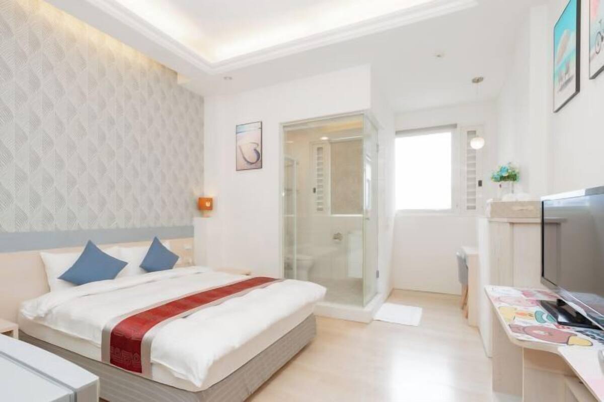 A modern hotel room with a bed, a desk with a computer, a TV, and a glass-enclosed shower area.