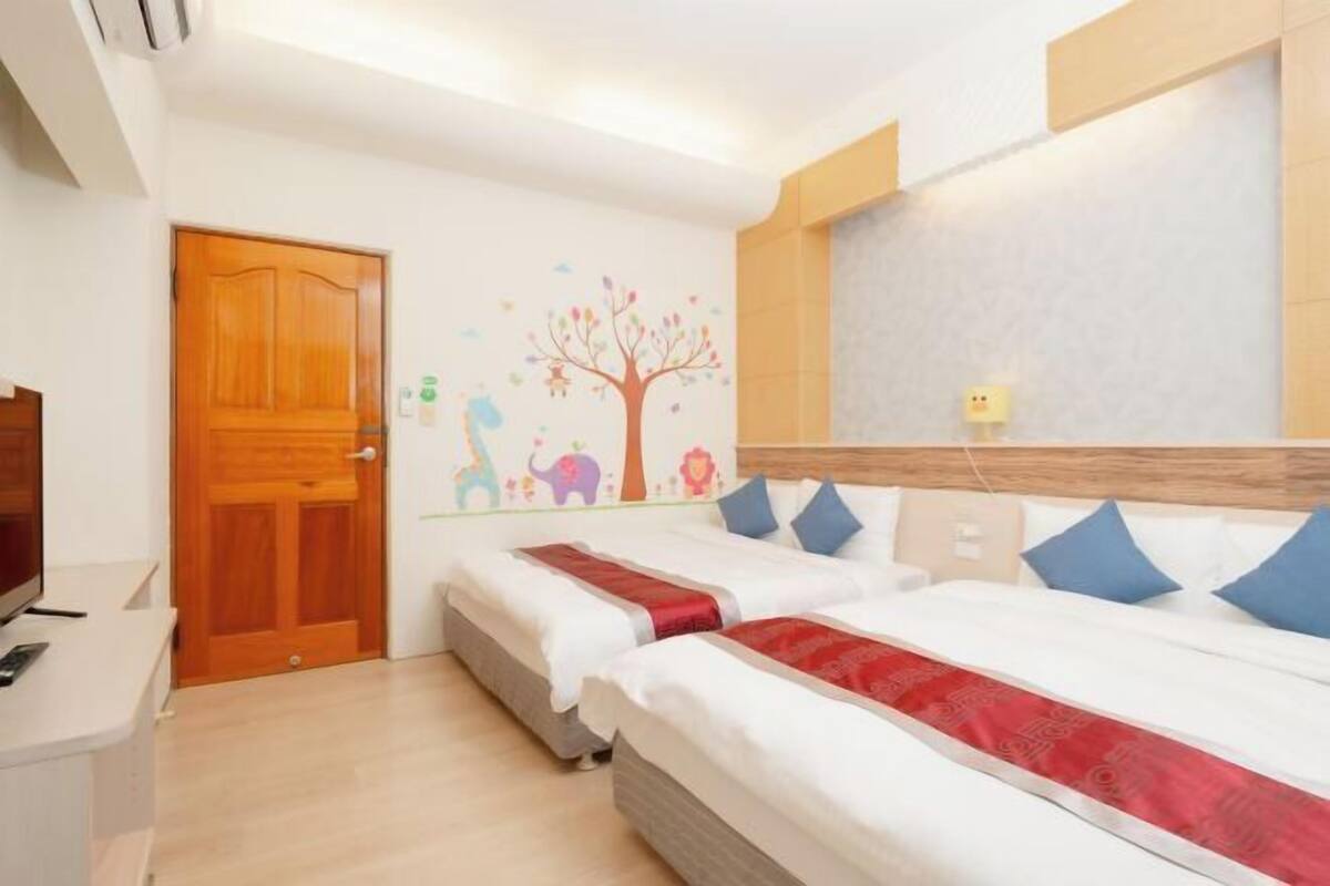 A hotel room with two beds, a wooden door, a wall with a colorful mural, and a small desk with a television.