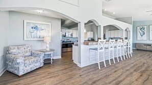 A spacious living area with a sofa and chairs, a kitchen in the background, and a dining area with a long table and chairs.