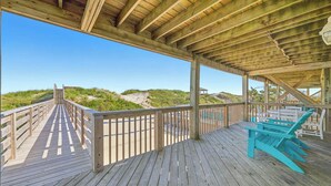 A wooden deck with a view of dunes and beach, featuring a covered seating area with chairs.