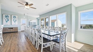 A spacious dining area with a long table and white chairs, large windows, and a ceiling fan.