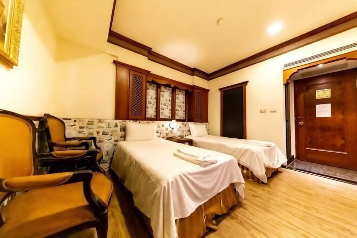 A hotel room with two beds, a wooden chair, a desk, and a door with a sign.