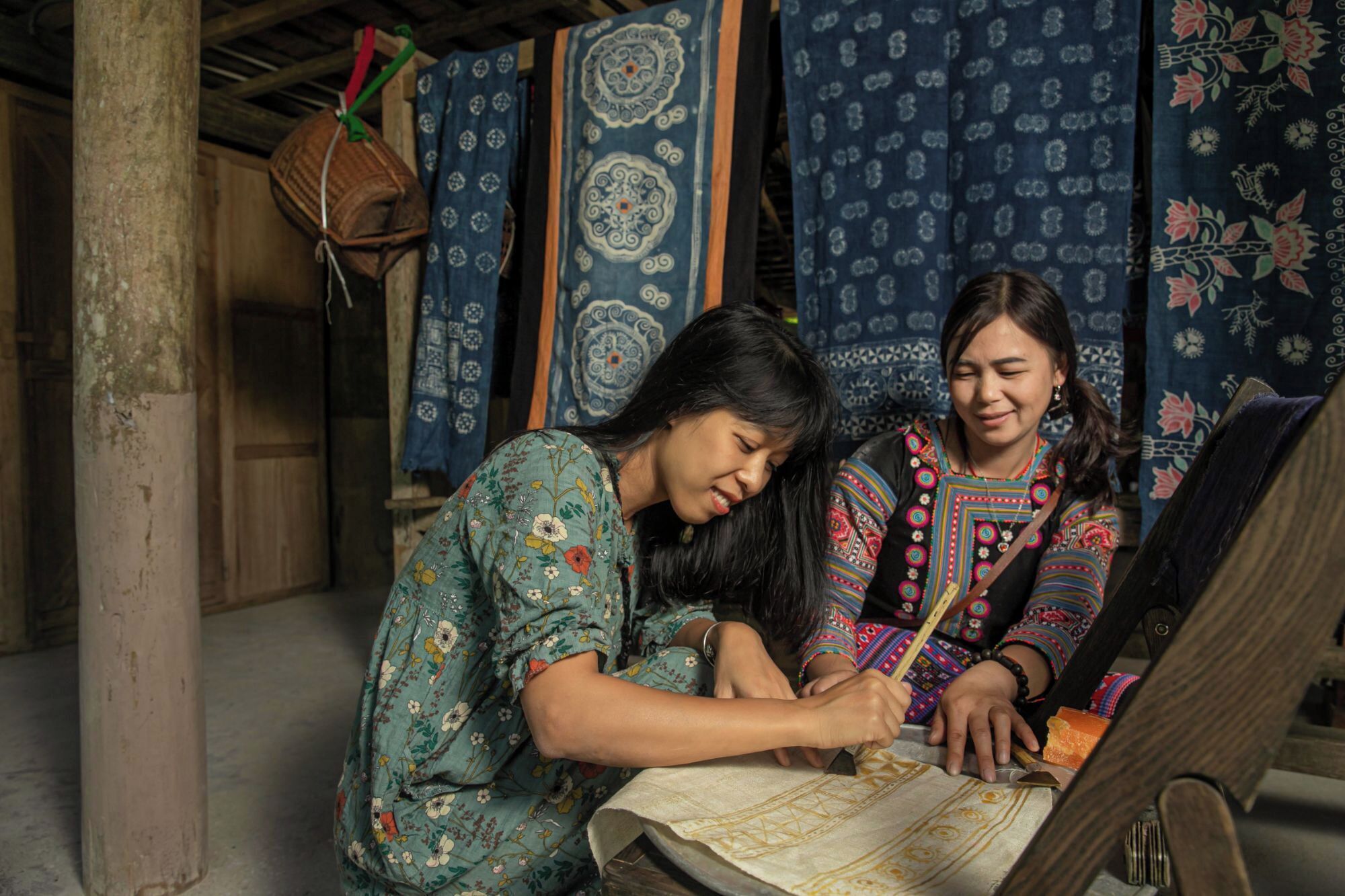 Two individuals working on a textile with intricate patterns, in a rustic setting with wooden structures and hanging fabrics.