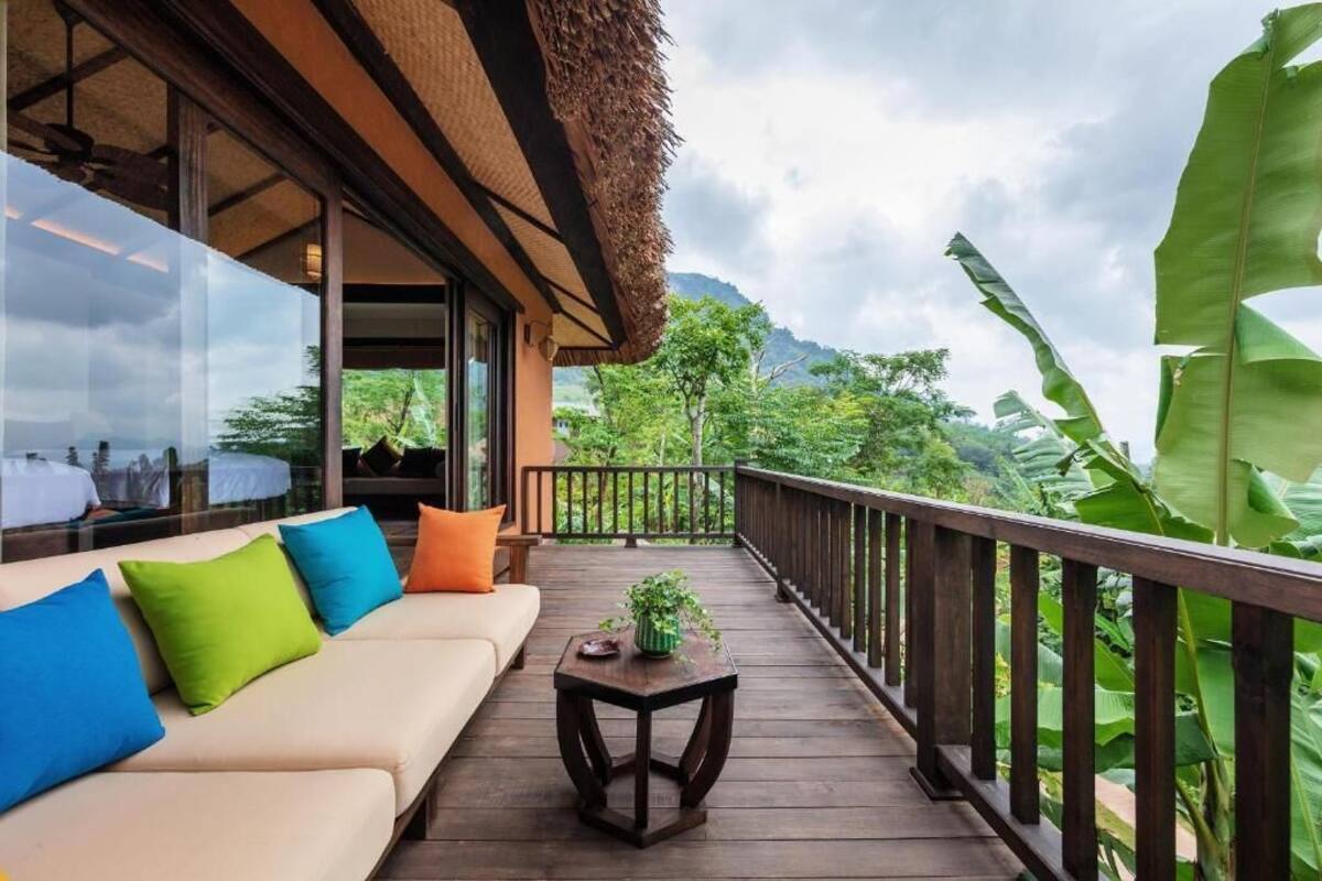 Una terraza de madera con un sofá y una mesita, con vistas a exuberante vegetación y montañas.