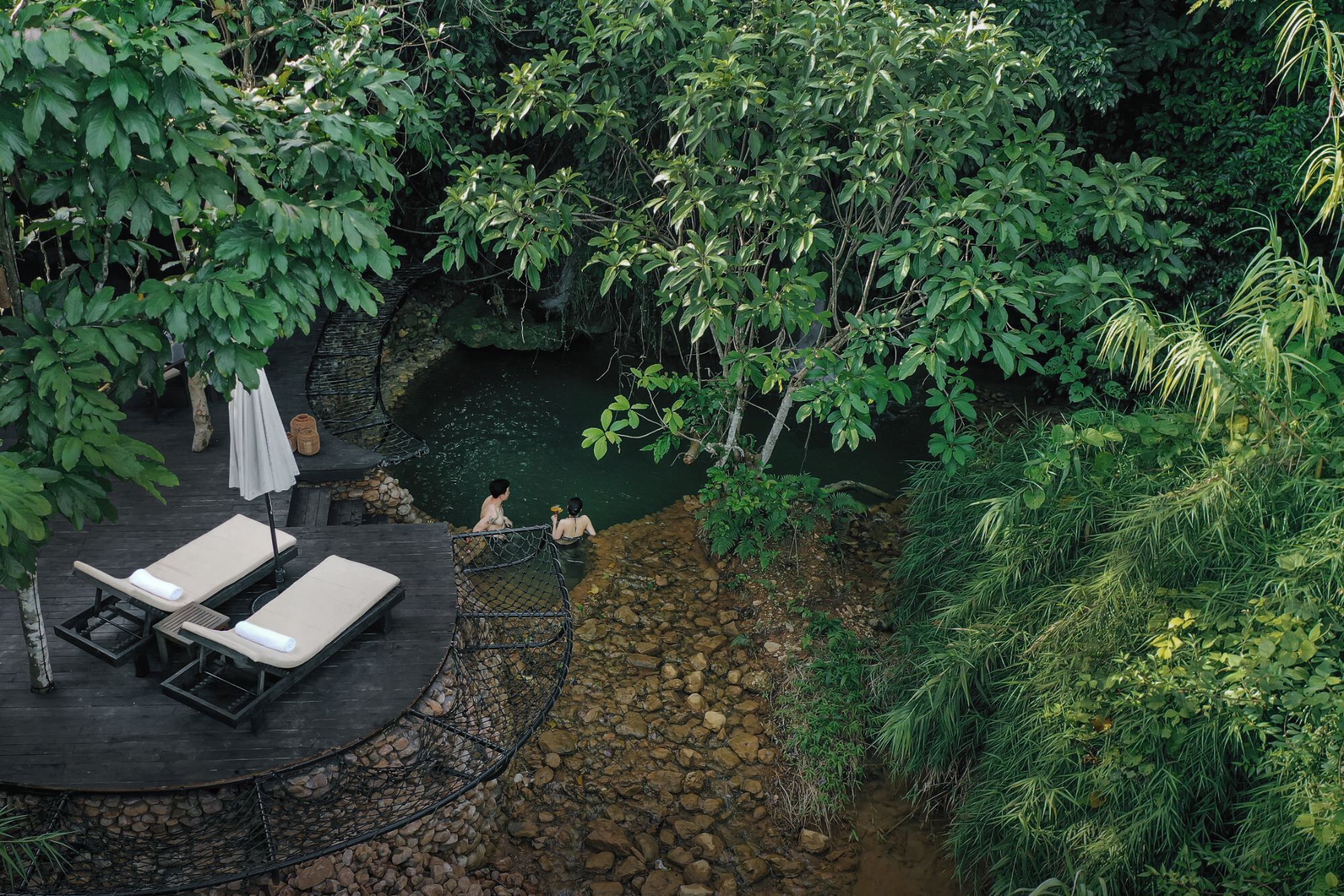 A secluded outdoor area with a pool surrounded by lush greenery, a wooden deck with lounge chairs, and a stone wall.
