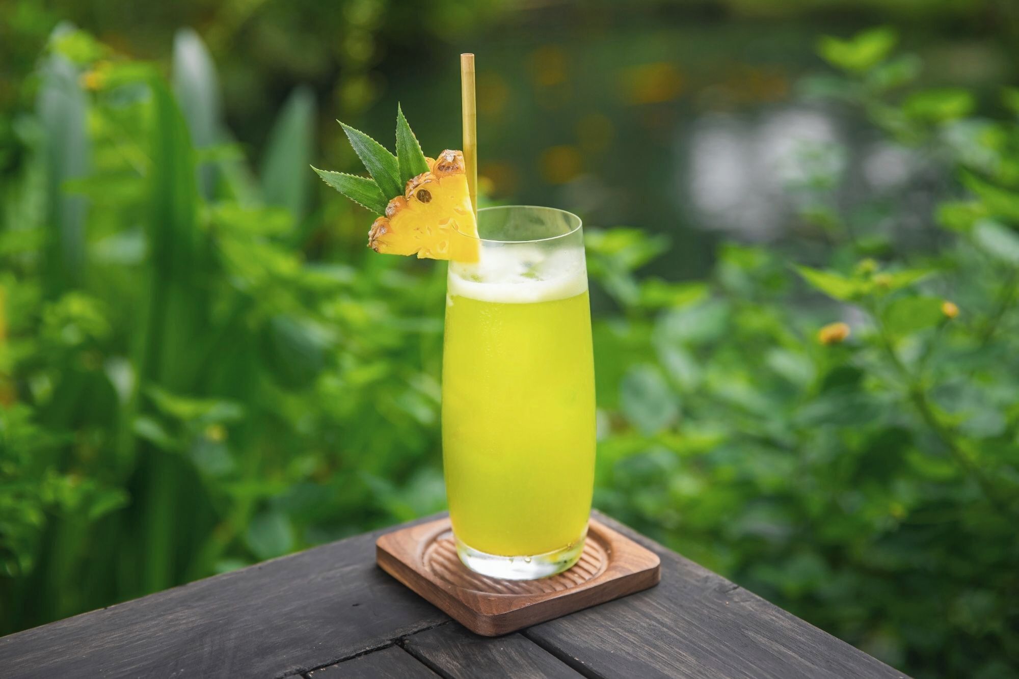 A tall glass with a yellow beverage, a pineapple slice, and a straw on a wooden coaster.