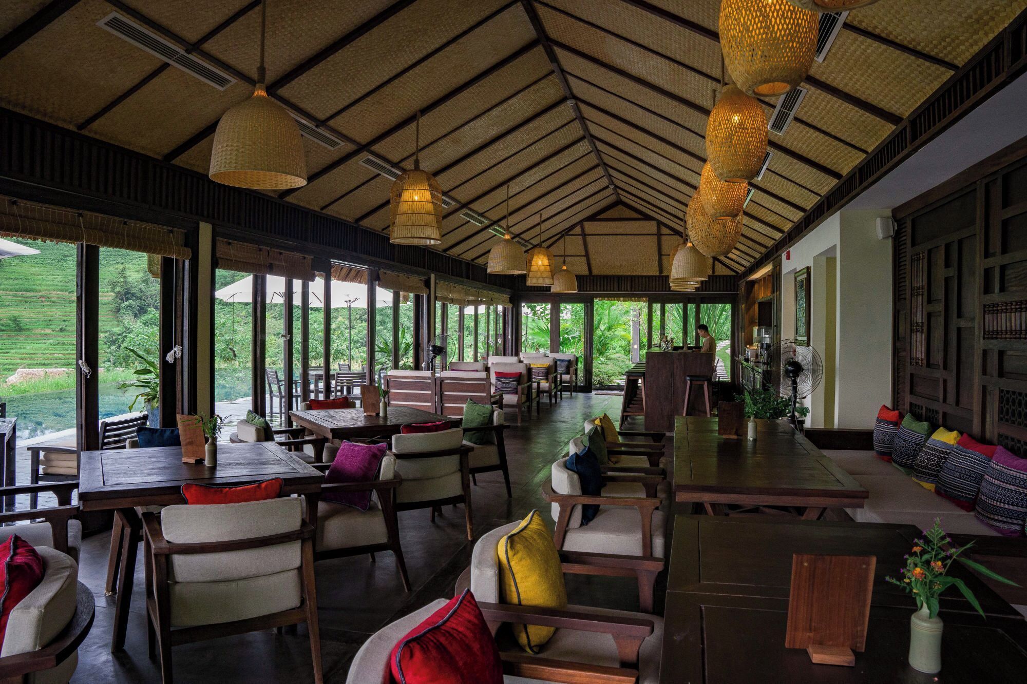Een ruime binnenruimte met houten tafels en stoelen, grote ramen met uitzicht op groen, en hangende geweven lampen.