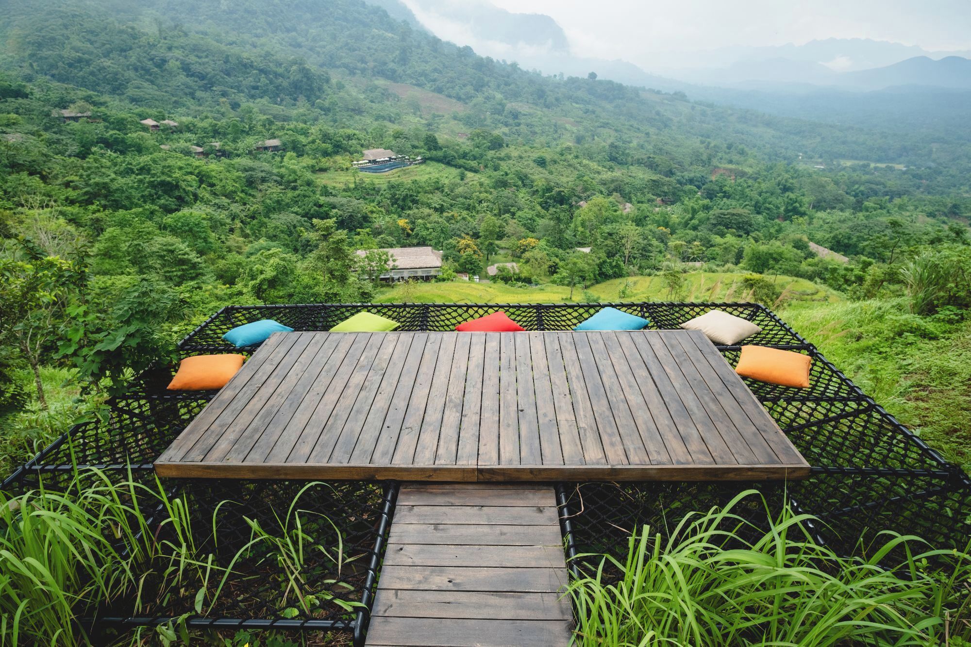 Una terraza de madera con cojines coloridos que da a un exuberante valle verde con edificios a lo lejos.