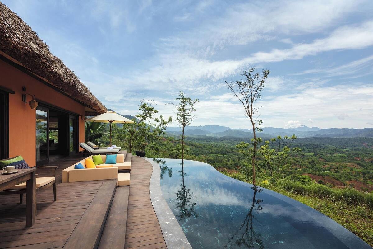Un moderno espacio lounge al aire libre con terraza de madera, asientos acolchados y una piscina infinita de cristal con vistas a un paisaje montañoso.