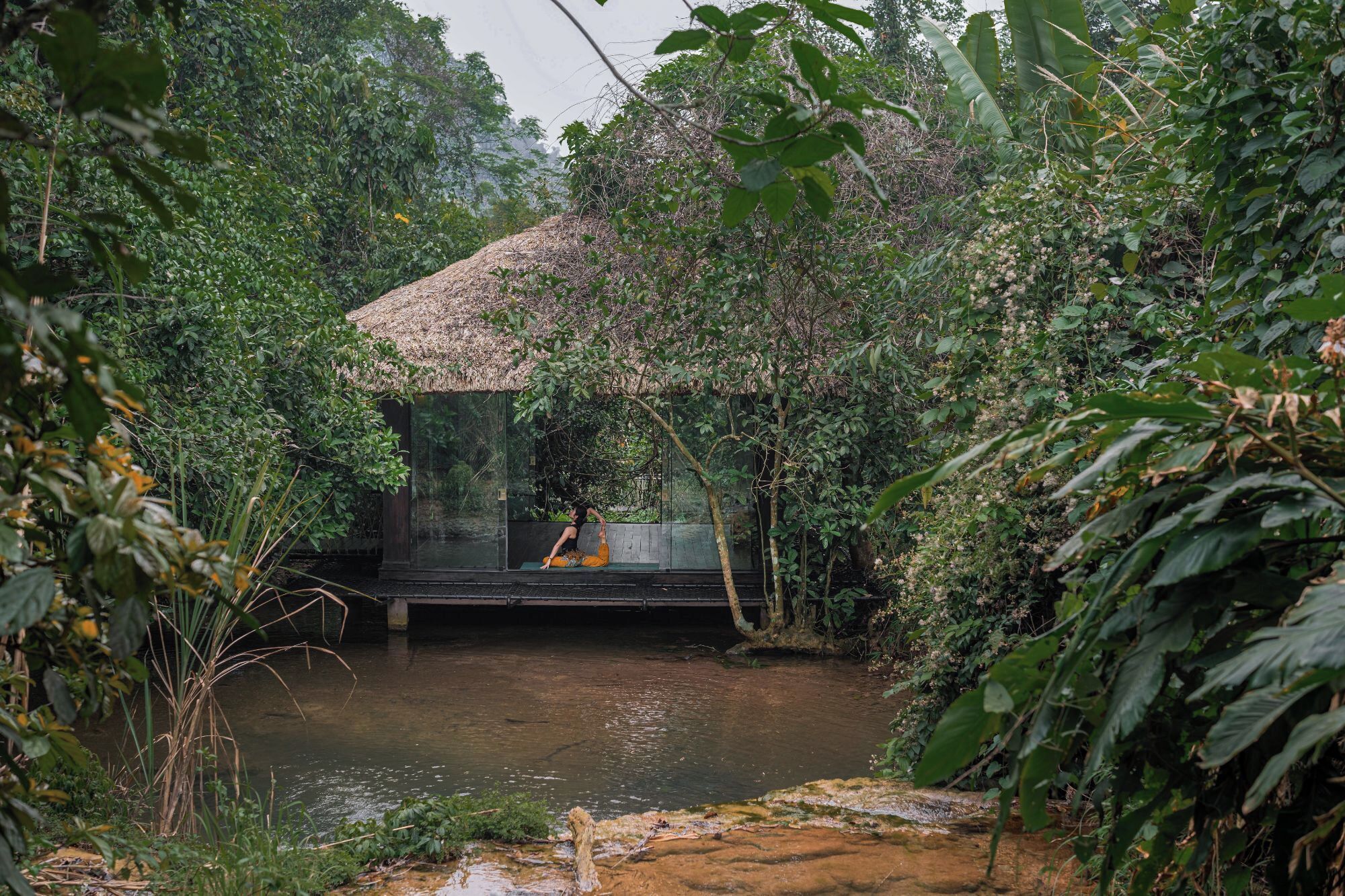Una estructura de madera aislada con un cerramiento de vidrio que da a un río tranquilo, rodeada de abundante vegetación.