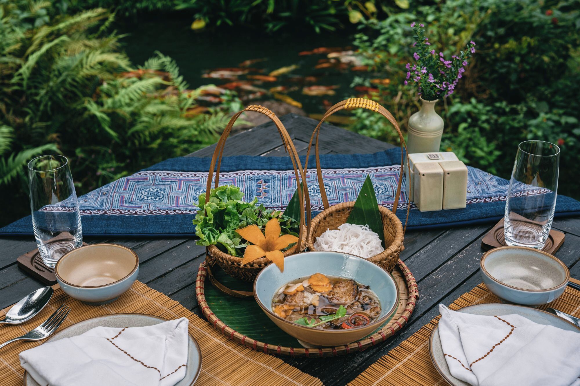 A table set with a meal, including a bowl of soup, a plate with a salad, and a basket of vegetables, with a view of a pond and greenery in the background.