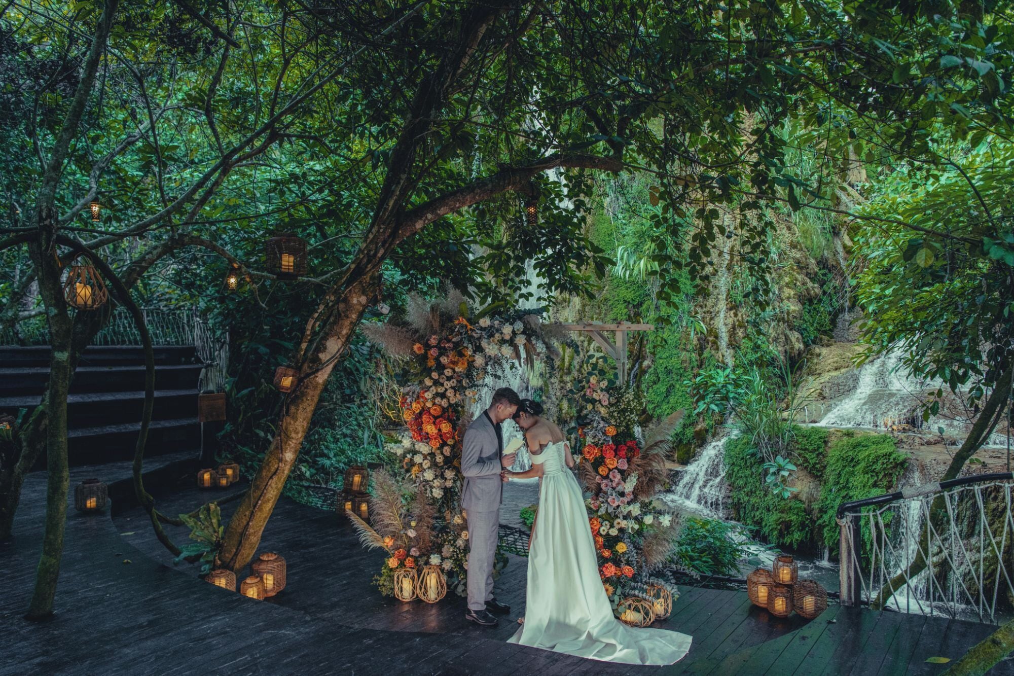 A wedding ceremony in a forested area with a wooden arch decorated with flowers and lanterns.