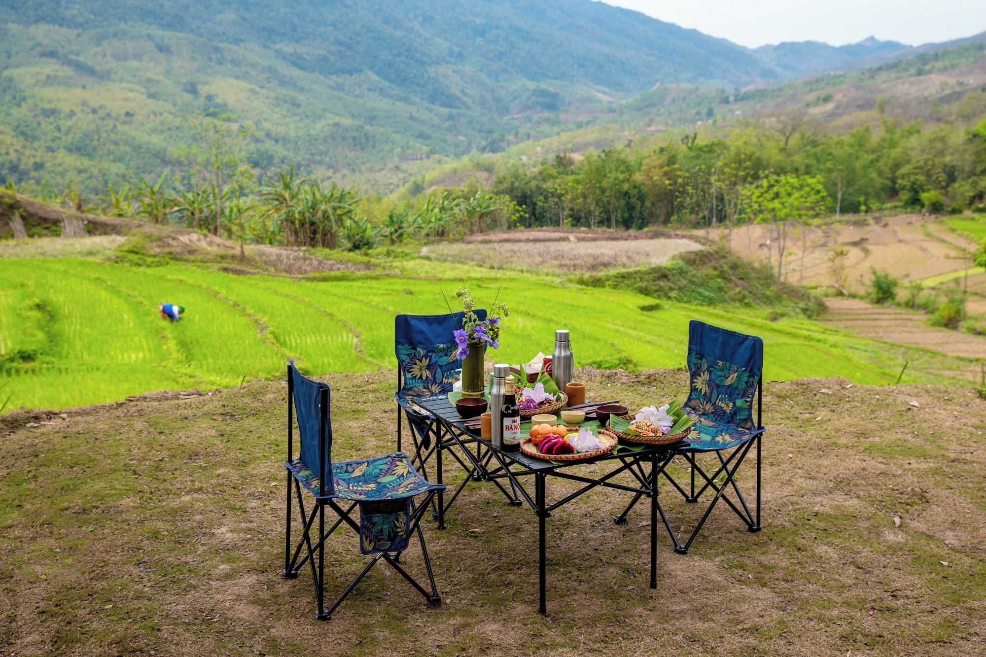 A dining table with chairs set up outdoors, surrounded by a lush green landscape and hills in the background.