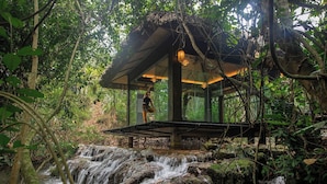 A wooden structure with a glass enclosure, surrounded by lush greenery and a cascading waterfall. - Avana Retreat (Bao La)