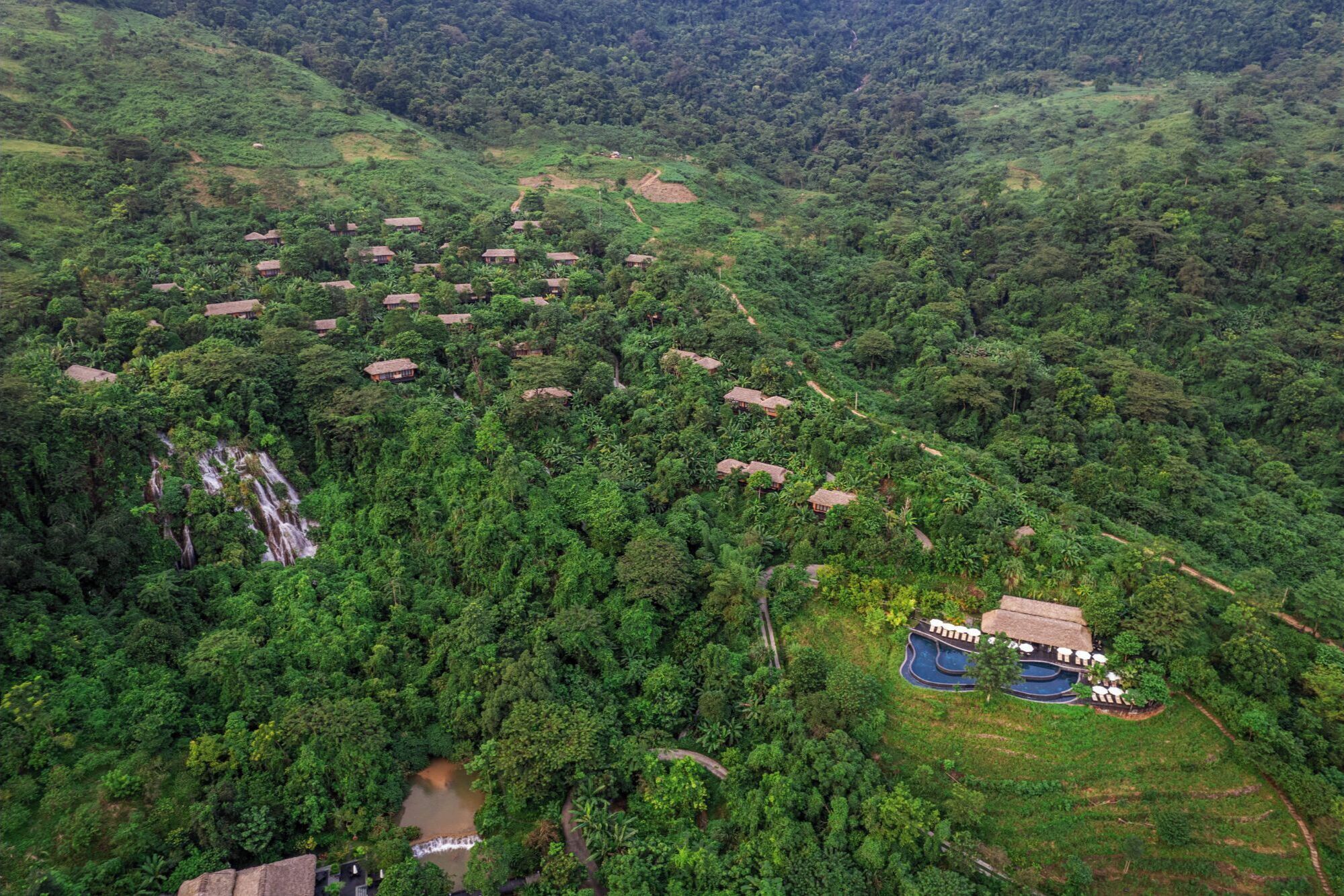 Vista aérea de un resort con varias cabañas rodeadas de un bosque denso, un pequeño área de piscina y una cascada.