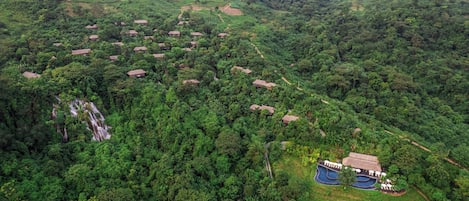 Vista aérea de un resort con varias cabañas rodeadas de un bosque denso, un pequeño área de piscina y una cascada.