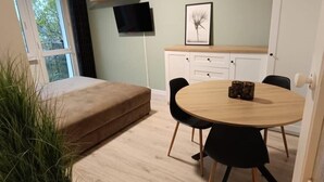 Un salon moderne avec un canapé, une table ronde en bois et des chaises noires. Un téléviseur est fixé au mur, il y a un tableau encadré et une petite étagère avec des plantes.