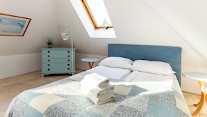 Ein Schlafzimmer mit schrägen Wänden, einem Bett mit blauer Kopfteil, einem kleinen Nachttisch, einer Kommode und einem Dachfenster.